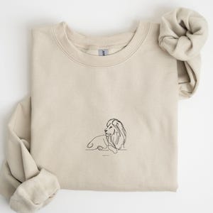 May include: A beige sweatshirt featuring a minimalist black line drawing of a lion in a resting pose. The text "John 11:1" is below the lion. The sweatshirt has a crew neck and long sleeves, with a matching scrunchie.