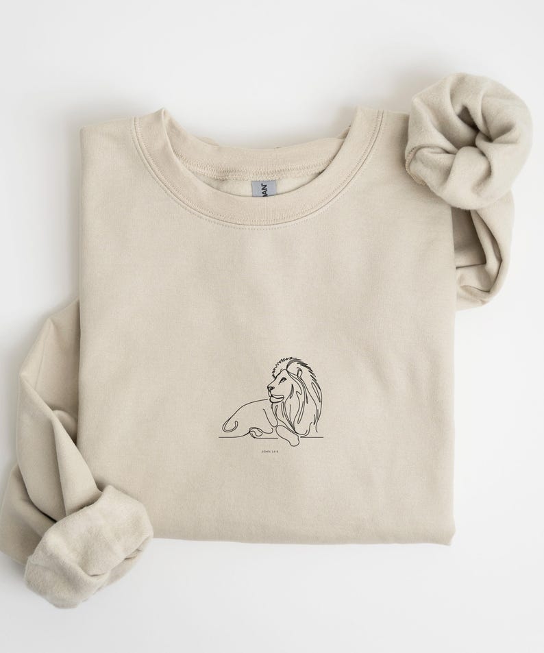 May include: A beige sweatshirt featuring a minimalist black line drawing of a lion in a resting pose. The text "John 11:1" is below the lion. The sweatshirt has a crew neck and long sleeves, with a matching scrunchie.