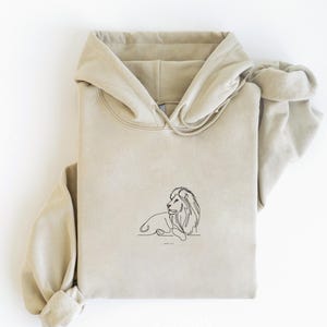 May include: A beige hoodie with a drawstring hood and a kangaroo pocket. The front of the hoodie features a minimalist black line drawing of a lion. The text "Jeremiah 1:5" is below the lion.