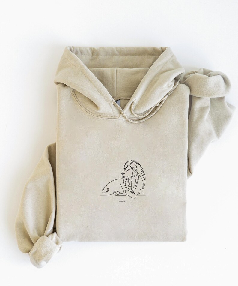 May include: A beige hoodie with a drawstring hood and a kangaroo pocket. The front of the hoodie features a minimalist black line drawing of a lion. The text "Jeremiah 1:5" is below the lion.