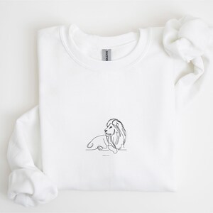 May include: White sweatshirt featuring a minimalist line drawing of a lion. The black outline depicts the lion in a resting pose, with the text "JOHN 11:9" below the image. The sweatshirt is made of a soft material.