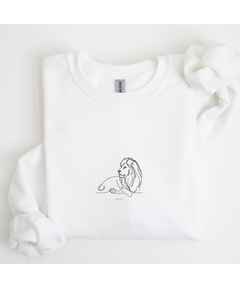 May include: White sweatshirt featuring a minimalist line drawing of a lion. The black outline depicts the lion in a resting pose, with the text "JOHN 11:9" below the image. The sweatshirt is made of a soft material.