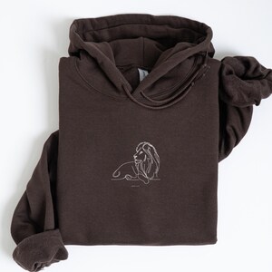 May include: A dark brown hoodie with a white line drawing of a lion on the front. The hoodie has a drawstring hood and long sleeves. The lion illustration is simple and elegant, with the words "GOOD VIBES" below it.