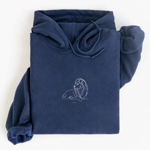 May include: A navy blue hoodie with a white line drawing of a lion on the front. The hoodie has a drawstring hood and long sleeves. The lion design is simple and elegant, with the text "JOHN 1:12" below it.