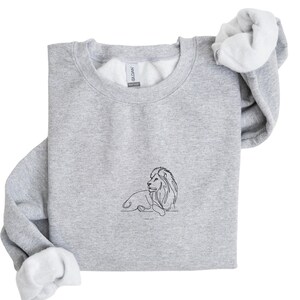 May include: A light gray sweatshirt with a crew neck and white interior. A black line drawing of a lion is centered on the front. The sweatshirt's sleeves are folded over, revealing the white lining. The brand name "Gildan" is visible on the tag.