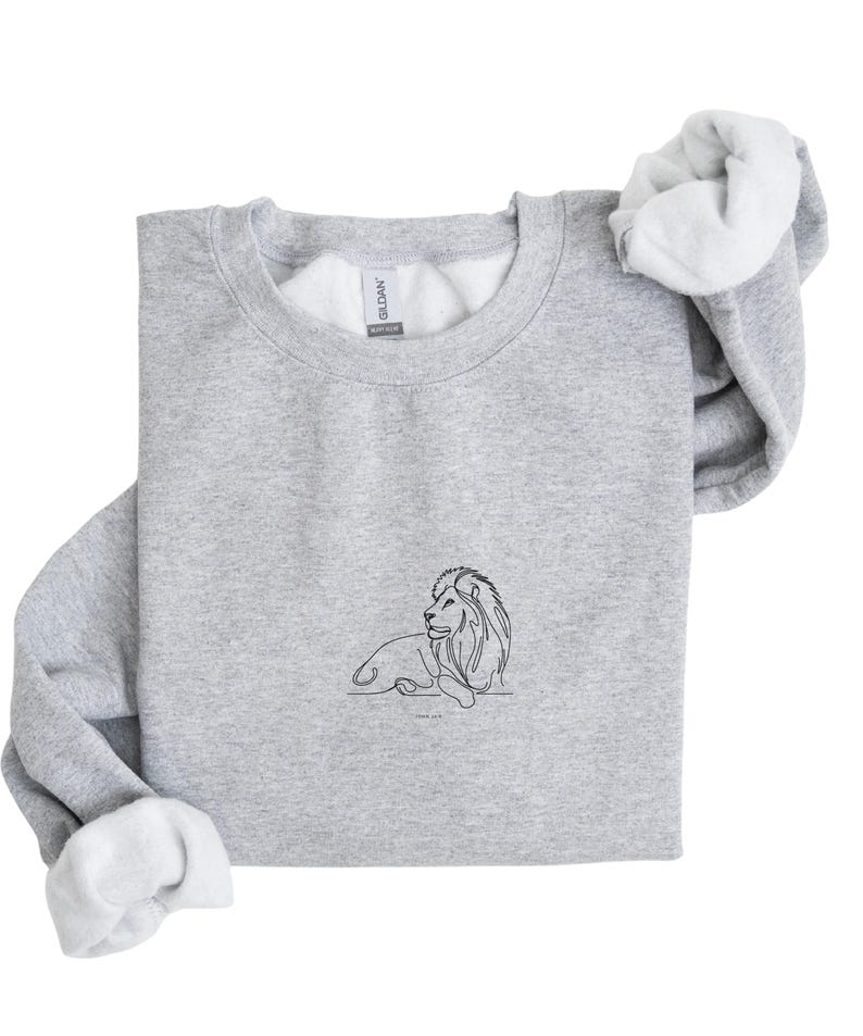 May include: A light gray sweatshirt with a crew neck and white interior. A black line drawing of a lion is centered on the front. The sweatshirt's sleeves are folded over, revealing the white lining. The brand name "Gildan" is visible on the tag.