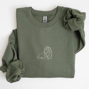 May include: Olive green sweatshirt with a white line drawing of a lion. The crew neck sweatshirt has long sleeves and a relaxed fit. The lion illustration is on the lower right side of the shirt.