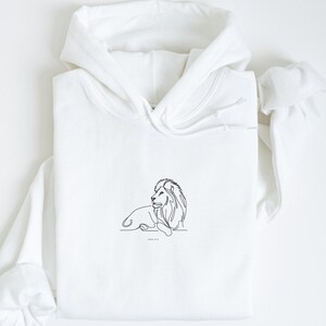 May include: White hoodie featuring a minimalist line drawing of a lion in black ink. The lion is depicted in a relaxed pose, with the text "JOHN 14:19" below the image. The hoodie has a drawstring hood and long sleeves.