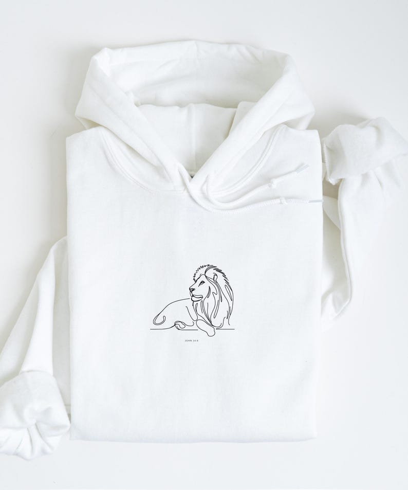 May include: White hoodie featuring a minimalist line drawing of a lion in black ink. The lion is depicted in a relaxed pose, with the text "JOHN 14:19" below the image. The hoodie has a drawstring hood and long sleeves.