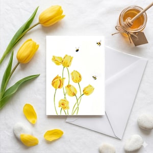 Könnte beinhalten: Eine weiße Grußkarte mit einem Aquarellgemälde von gelben Tulpen und drei Bienen. Die Karte wird von einem weißen Umschlag, einem Honigglas mit einem Holzstäbchen und Blütenblättern und Steinen auf weißer Oberfläche begleitet.
