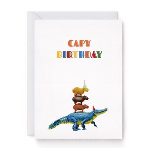 May include: A white birthday card with the text "CAPY BIRTHDAY" in bright colours. The card shows a blue alligator with capybaras stacked on top, each wearing a party hat, creating a fun and quirky design.