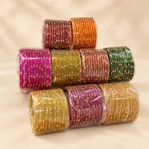Peut inclure: Une collection de bracelets joncs en verre multicolores disposés en piles et enveloppés dans du plastique transparent. Les bracelets présentent une finition translucide avec de petites paillettes dorées, déclinés en rouge, orange, rose, or et vert.