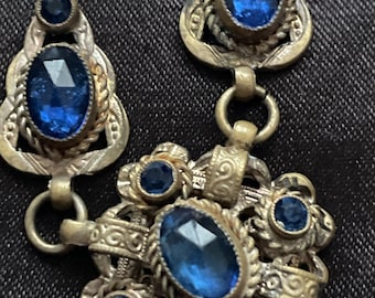 Op de afbeelding: Close-up van een vintage ketting met sierlijk filigraan metaalwerk. De ketting heeft grote, ovale, gefacetteerde blauwe edelstenen en kleinere ronde blauwe stenen. Het metaal heeft een aangetaste, antieke uitstraling, met ingewikkelde details.