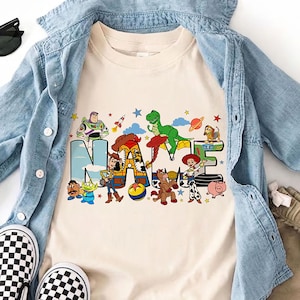 Puede incluir: Una camiseta color crema con la palabra "NAME" en un dise&ntilde;o colorido con personajes de la pel&iacute;cula Toy Story. La camiseta se combina con una camisa vaquera azul claro, pantalones cortos color caqui y zapatos sin cordones a cuadros blancos y negros. Tambi&eacute;n se incluyen gafas de sol.