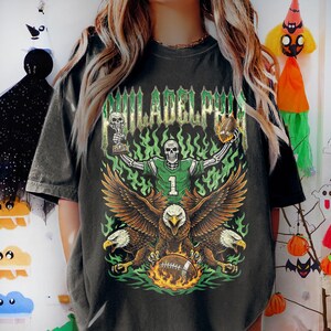 May include: Dark gray t-shirt with a graphic featuring a skeleton in a football uniform, holding a trophy and a football. The design includes an eagle with a flaming football, green flames, and the word "PHILADELPHIA".