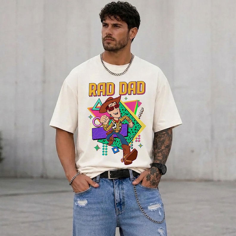 May include: Cream-colored t-shirt with the text "RAD DAD" in a retro font. The shirt features a cartoon illustration of a cowboy character. The design includes geometric shapes in various colors.
