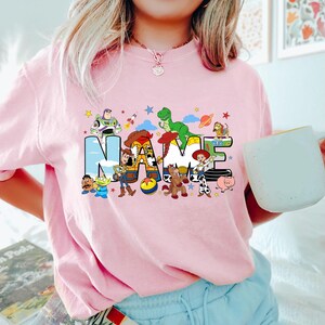 Puede incluir: Camiseta rosa claro con la palabra "NAME" en letras grandes y coloridas, con personajes de dibujos animados. El dise&ntilde;o incluye un cohete, estrellas y planetas. La camiseta es de color rosa claro.