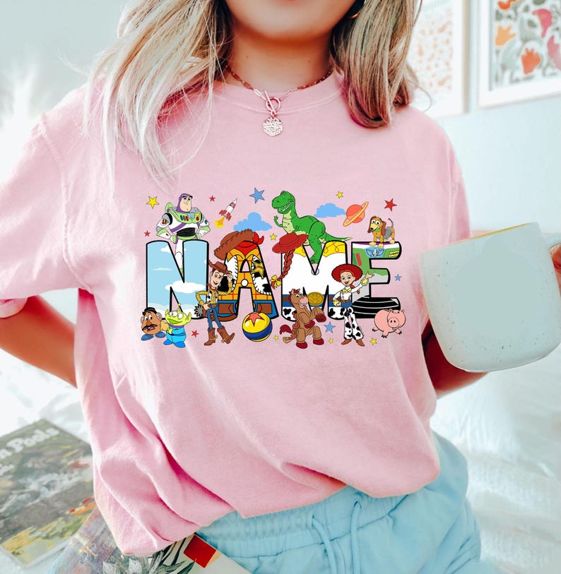 Puede incluir: Camiseta rosa claro con la palabra "NAME" en letras grandes y coloridas, con personajes de dibujos animados. El dise&ntilde;o incluye un cohete, estrellas y planetas. La camiseta es de color rosa claro.