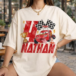 May include: Cream-colored t-shirt featuring a red graphic with the number 4, a trophy, and a cartoon car with checkered flags. The name "NATHAN" is printed in red below the number. The design has a watercolor effect.