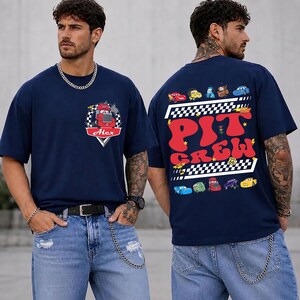 Op de afbeelding: Een marineblauw t-shirt met korte mouwen en een kleine afbeelding van een rode vrachtwagen op de linkerborst met de naam Alex. De achterkant toont een grote afbeelding met de tekst PIT CREW, geblokte patronen en diverse kleurrijke tekenfilmauto's.