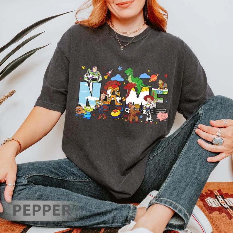 Puede incluir: Camiseta gris oscuro con la palabra "NAME" en letras coloridas, cada letra decorada con personajes de una popular pel&iacute;cula de animaci&oacute;n. La camiseta es de un estilo c&oacute;modo e informal.