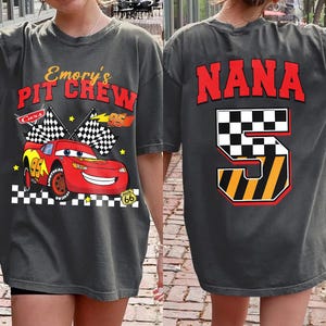 May include: Dark gray t-shirt featuring a red race car with the text "Emory's Pit Crew" and checkered flags. The back of the shirt displays the word "NANA" in red above a checkered number 5.