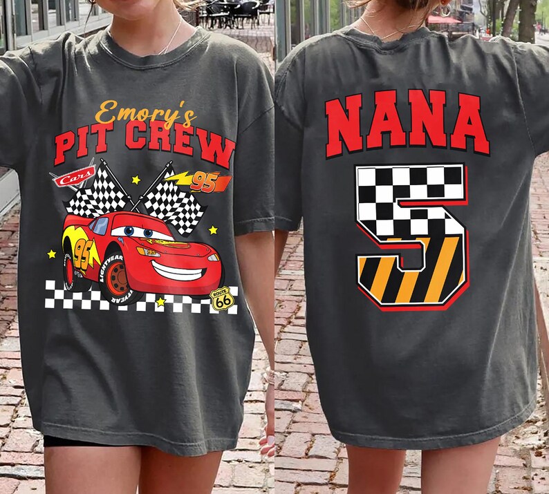 May include: Dark gray t-shirt featuring a red race car with the text "Emory's Pit Crew" and checkered flags. The back of the shirt displays the word "NANA" in red above a checkered number 5.