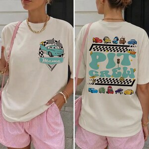 Op de afbeelding: Een off-white t-shirt met een kleine afbeelding van een lichtblauwe auto op de borst met de tekst "Meme". De achterkant toont een grote "PIT CREW"-afbeelding in lichtblauw met diverse cartoonauto's, geblokte patronen en een Route 66-bord.