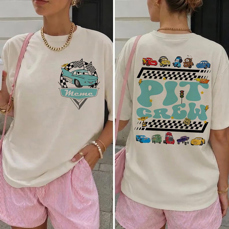 Op de afbeelding: Een off-white t-shirt met een kleine afbeelding van een lichtblauwe auto op de borst met de tekst "Meme". De achterkant toont een grote "PIT CREW"-afbeelding in lichtblauw met diverse cartoonauto's, geblokte patronen en een Route 66-bord.