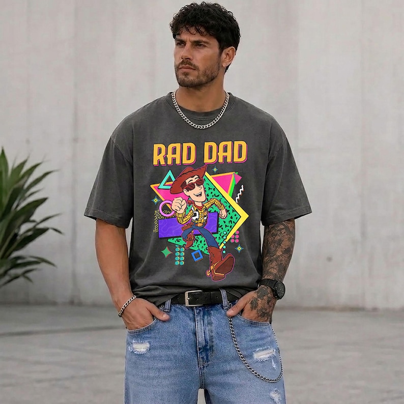 May include: Dark gray t-shirt with the text "RAD DAD" and a cartoon graphic of a cowboy character. The graphic features a colorful geometric design. The shirt is worn with blue jeans and a black belt.
