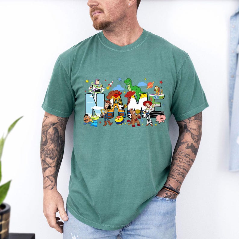 Puede incluir: Camiseta verde azulado con la palabra "NAME" en una fuente colorida y juguetona, decorada con personajes de dibujos animados de una popular pel&iacute;cula de animaci&oacute;n. El dise&ntilde;o incluye personajes como Buzz Lightyear, Woody y Rex.