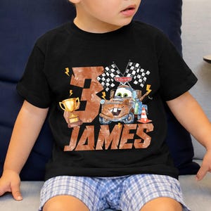 May include: Black t-shirt featuring a watercolor design with the number "3", a gold trophy, and the name "JAMES". The design includes a cartoon car character with checkered flags and lightning bolts.
