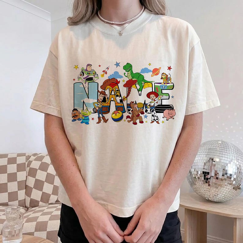 Puede incluir: Camiseta color crema con la palabra "NAME" en una fuente colorida y caricaturesca, con varios personajes de Toy Story incorporados en las letras. El dise&ntilde;o incluye a Buzz Lightyear, Woody y Rex. La camiseta tiene mangas cortas.