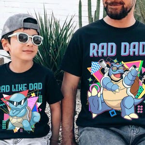 Puede incluir: Camisetas negras con gráficos a juego. Una camiseta dice "RAD DAD" y presenta un personaje de dibujos animados azul con gafas de sol. La otra camiseta dice "RAD LIKE DAD" y tiene un diseño similar. Ambas camisetas tienen un estilo retro de los años 80.