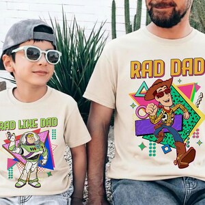 May include: Two beige t-shirts with cartoon graphics. One says "RAD DAD" with a Woody character, the other says "RAD LIKE DAD" with a Buzz Lightyear character. Both shirts have a retro 80s design.