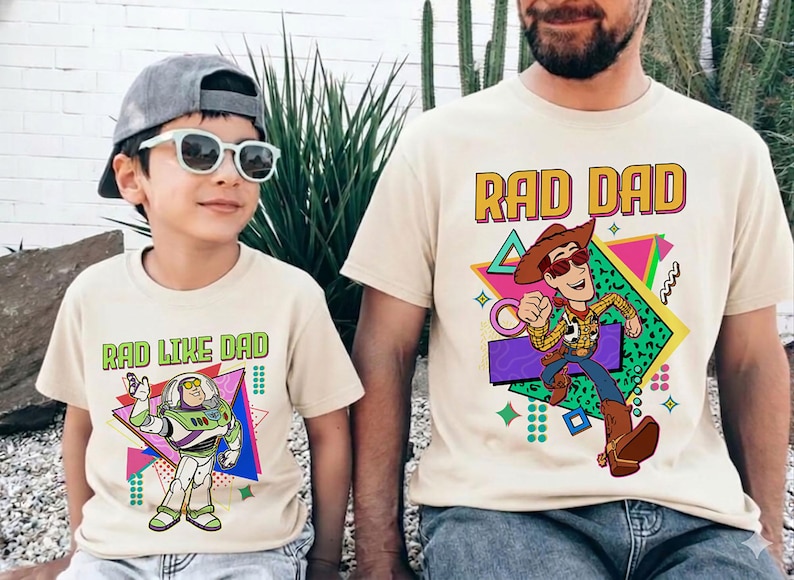 May include: Two beige t-shirts with cartoon graphics. One says "RAD DAD" with a Woody character, the other says "RAD LIKE DAD" with a Buzz Lightyear character. Both shirts have a retro 80s design.