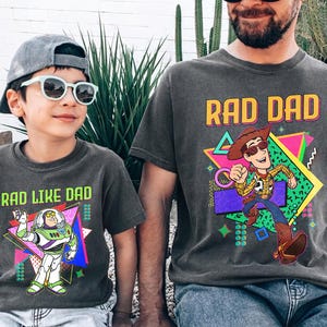 May include: Two matching charcoal gray t-shirts. One says "RAD DAD" with a cartoon Woody character. The other says "RAD LIKE DAD" with a Buzz Lightyear graphic. Both shirts have a retro 80s design.