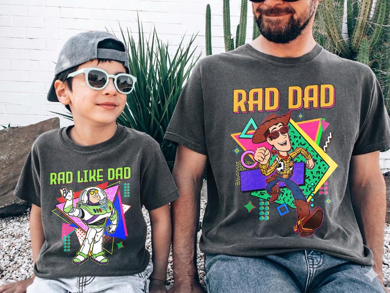 May include: Two matching charcoal gray t-shirts. One says "RAD DAD" with a cartoon Woody character. The other says "RAD LIKE DAD" with a Buzz Lightyear graphic. Both shirts have a retro 80s design.