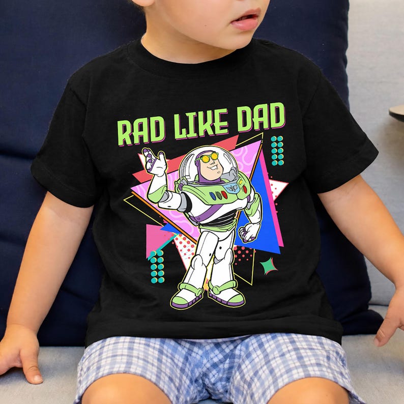 May include: Black t-shirt featuring a graphic of Buzz Lightyear with the text "RAD LIKE DAD" in neon green. The design includes a colorful geometric background. The shirt is worn by a child.