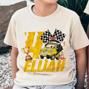 May include: A beige t-shirt featuring a yellow cartoon car with the text "Cars" on the roof, checkered flags, a trophy, and the name "ELIJAH". The number "4" is prominently displayed in a yellow, distressed font.