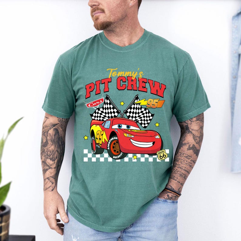 May include: A teal green t-shirt featuring a graphic of Lightning McQueen from the movie Cars. The design includes the text "Tommy's Pit Crew" and checkered flags. The number 95 is prominently displayed on the car.