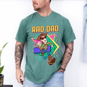 May include: Green t-shirt with the text "RAD DAD" in a colorful 80s style font. The shirt features a cartoon cowboy character wearing sunglasses and a cowboy hat. The character is running with a gun holster.