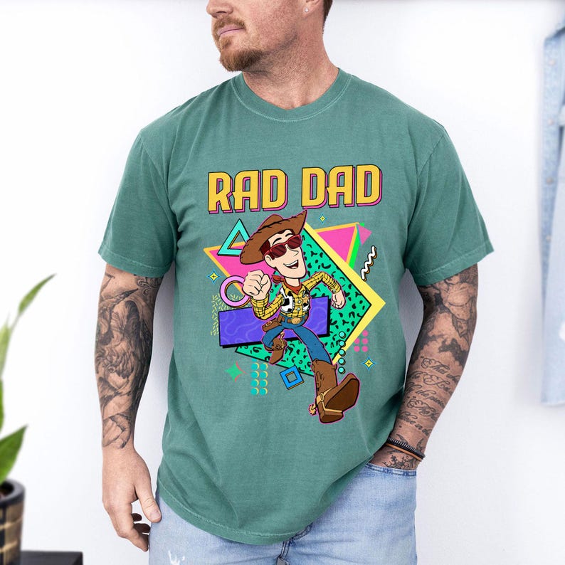 May include: Green t-shirt with the text "RAD DAD" in a colorful 80s style font. The shirt features a cartoon cowboy character wearing sunglasses and a cowboy hat. The character is running with a gun holster.
