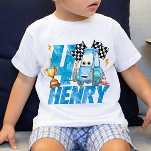 May include: White t-shirt with a blue and green cartoon car graphic, checkered flags, and a gold trophy. The number "4" and the name "HENRY" are printed in blue. A fun birthday shirt for a child.