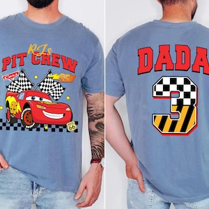 May include: A light blue t-shirt featuring a graphic of a red race car with the text "RJs Pit Crew Cars 95" on the front. The back of the shirt displays the word "DADA" above a number "3" with a checkered flag design.