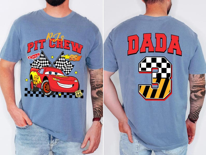 May include: A light blue t-shirt featuring a graphic of a red race car with the text "RJs Pit Crew Cars 95" on the front. The back of the shirt displays the word "DADA" above a number "3" with a checkered flag design.