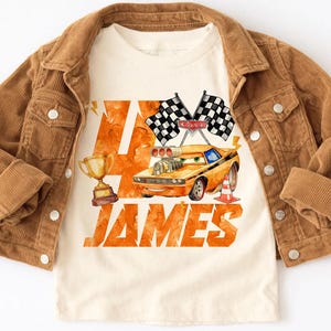 May include: A tan t-shirt featuring a cartoon race car design with the name "JAMES" in orange lettering. The shirt is paired with a brown corduroy jacket. The design includes checkered flags, a trophy, and a traffic cone.