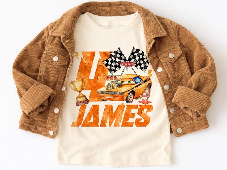 May include: A tan t-shirt featuring a cartoon race car design with the name "JAMES" in orange lettering. The shirt is paired with a brown corduroy jacket. The design includes checkered flags, a trophy, and a traffic cone.