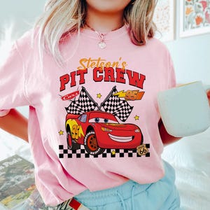 May include: A light pink crew neck t-shirt featuring a graphic of the character Lightning McQueen from the movie Cars. The graphic includes the text "Stetson's Pit Crew" and checkered flags. The number 95 is also featured.