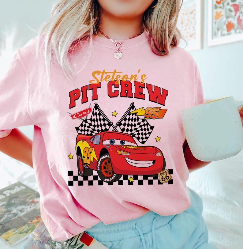 May include: A light pink crew neck t-shirt featuring a graphic of the character Lightning McQueen from the movie Cars. The graphic includes the text "Stetson's Pit Crew" and checkered flags. The number 95 is also featured.